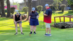 3 players stand on the green with one putting a shot into the hole