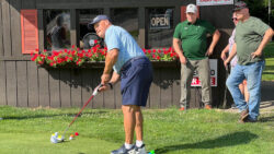 Player putts towards hole on putting green in front of the clubhouse while other players look on